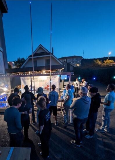 Foto: Besucherinnen und Besucher vor dem Thingware-Gebäude in der Dämmerung, im Hintergrund die hohen Masten der TWANG32-LED-Bahnen